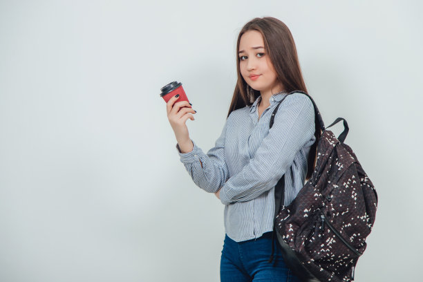 年轻美丽的一个学生女孩站在一个背包里拿着一杯咖啡。图片下载