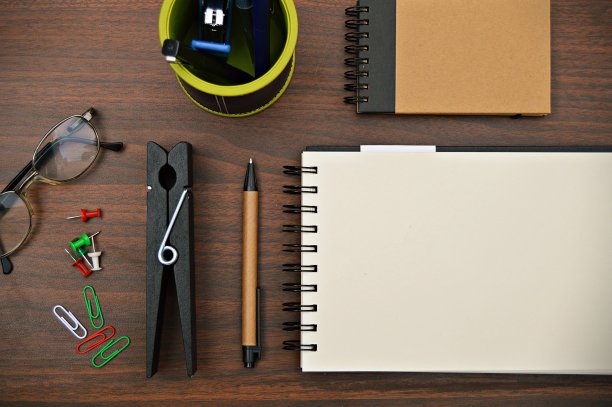 一个文具的横向照片，如钢笔，日记，笔架与笔，订书机，一个大回形针，回形针和别针放置在一个木制的深棕色水平背景上。图片下载