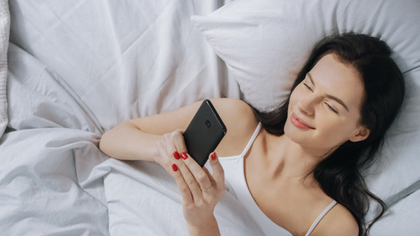 在她的床上用触屏智能手机和微笑，与朋友聊天，使用社交媒体，张贴图片，分享想法的美丽的黑发女人的肖像。自顶向下。图片下载