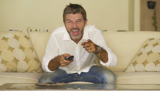 家庭生活肖像年轻的有吸引力的快乐和开朗的男人看电视有趣的节目或喜剧电影，笑着轻松地拿着电视遥控器坐在客厅沙发沙发图片下载