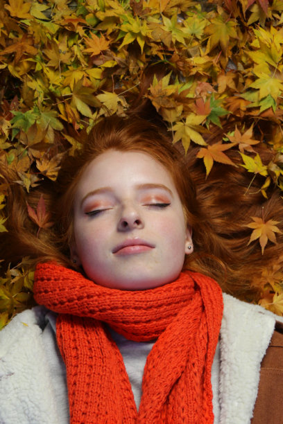 美丽的年轻漂亮的英国女孩的形象睡着了明亮的姜红色头发，长长的头发躺在黄橙色的秋天枫叶外面的秋天的颜色在花园在寒冷的秋天，毛皮麂皮夹克，手织橙色围巾图片下载