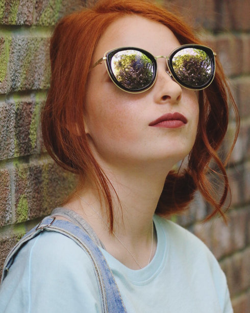 这是一个忧郁忧郁的少女的形象她的红头发向下看着，在涂鸦的砖墙前陷入了沉思，红头发穿着蓝色的牛仔工作服，戴着太阳镜，化妆，涂着深红色的口红，姜黄色的头发绑在后面，卷着图片下载