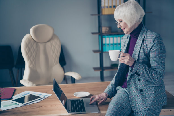 年老的商务女士的资料照片手热饮料杯沟通skype亲戚坐桌子办公室穿specs服装套装夹克图片下载