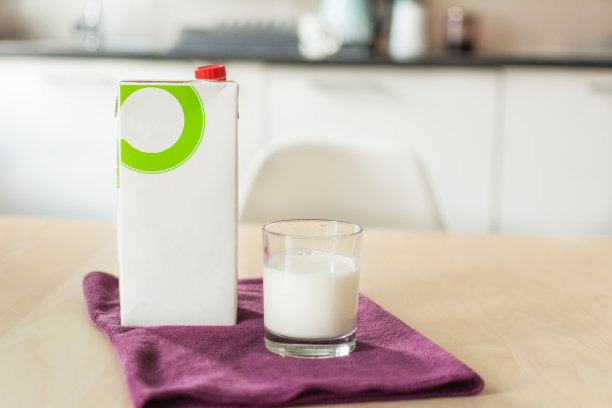 木桌上放着一杯牛奶、餐巾和一个纸瓶，厨房背景是模糊的白色。图片下载