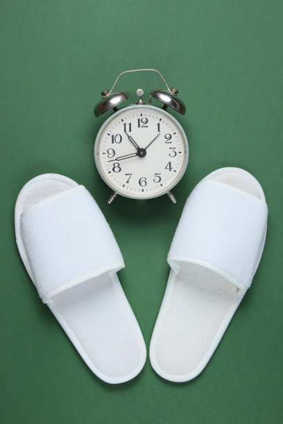 白色酒店睡鞋和闹钟在绿色背景。时间睡觉。俯视图图片下载