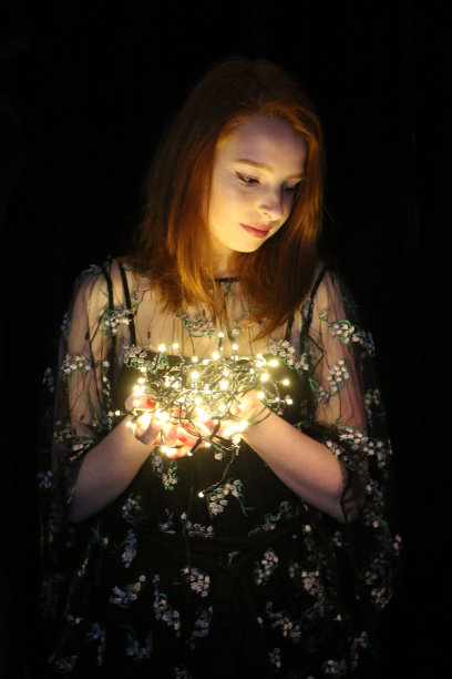 美丽的年轻女孩的形象，红发女人与圣诞树彩灯和长长的红姜头发在长鲍勃发型站在黑暗的房间晚上穿着漂亮的小黑裙LBD与鲜花刺绣，举着白色的彩灯照明图片下载
