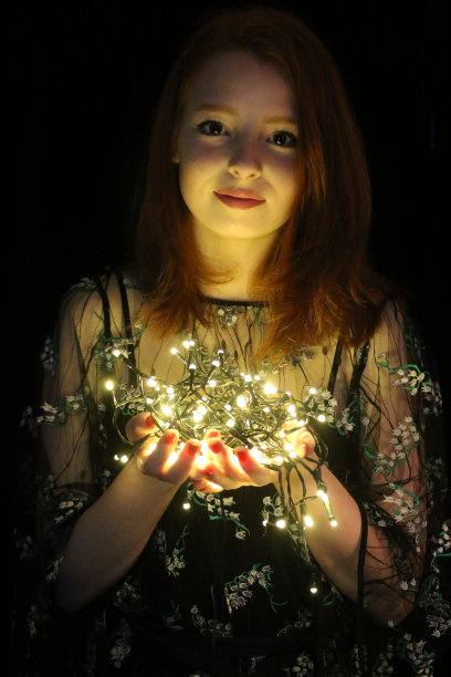 美丽的年轻女孩的形象，红发女人与圣诞树彩灯和长长的红姜头发在长鲍勃发型站在黑暗的房间晚上穿着漂亮的小黑裙LBD与鲜花刺绣，举着白色的彩灯照明图片下载