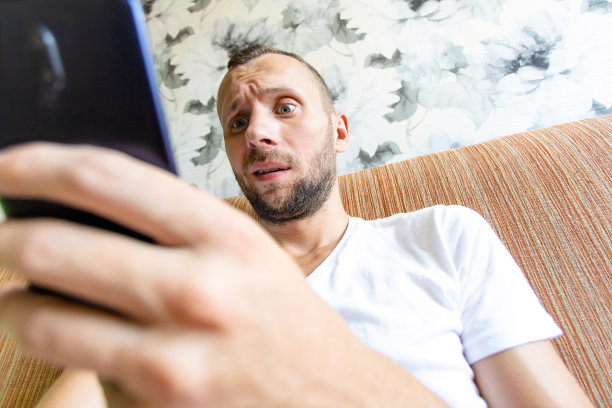 一个年轻人坐在沙发上，一脸惊讶地看着手机屏幕图片下载