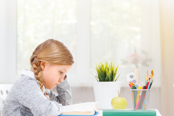 一个小学生女孩坐在桌子旁看书的侧视图。教育和学校的概念。阳光从窗户射进来。模糊的背景图片下载