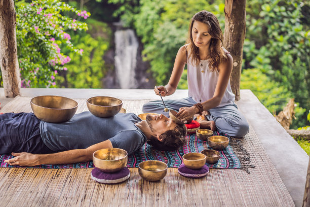 温泉沙龙的尼泊尔佛铜歌唱碗。年轻漂亮的男人做按摩治疗唱歌碗在温泉对着瀑布。健全的治疗，娱乐，冥想，健康的生活方式和身体护理理念图片下载