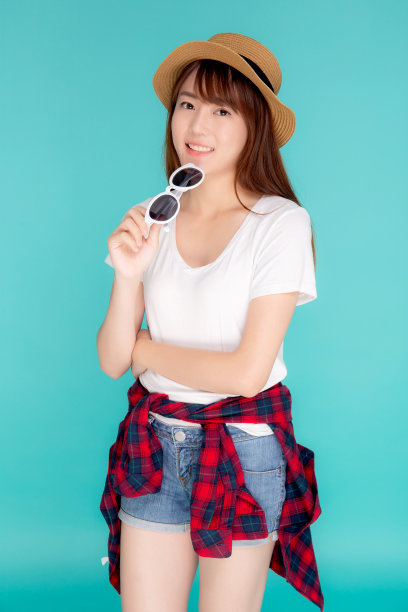 美丽的肖像年轻的亚洲女人戴着帽子和拿着太阳镜微笑表情自信享受夏天的假期孤立在蓝色的背景，女孩开朗穿着时尚的旅游概念。图片下载