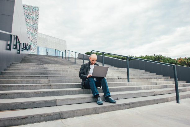 有魅力的成年成功秃顶胡子男人穿着黑色夹克在城市的楼梯上使用笔记本电脑图片下载