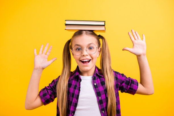 小女士小学生的特写照片，拿着书在头上笑，举起双臂，穿specs格子衬衫孤立的黄色背景图片下载