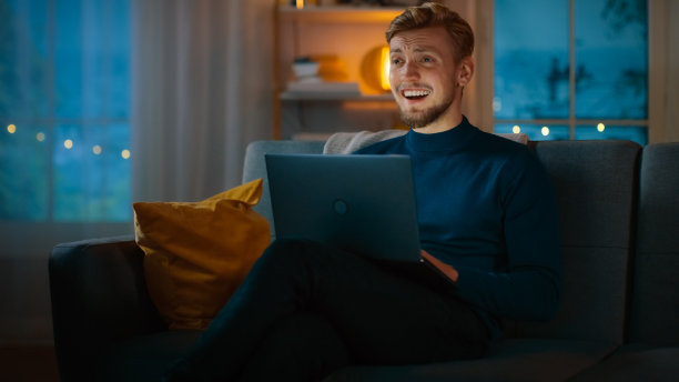 晚上，英俊的年轻人坐在沙发上用笔记本电脑工作，看电视喜剧和大笑。图片下载