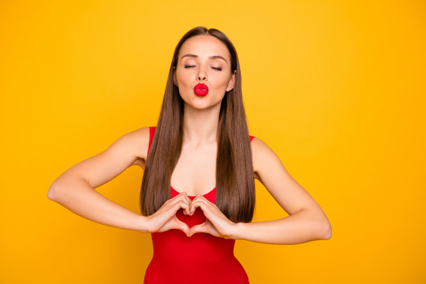 美丽的女士明亮的照片送飞吻红色的口红显示手指心形的图形浪漫的约会穿红色的泳衣孤立的黄色背景图片下载