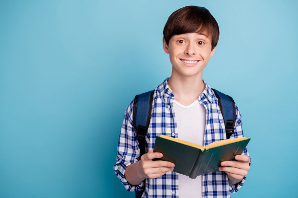 一个快乐的男孩的肖像，拿着印刷书，穿着格子衬衫，背包孤立在蓝色的背景图片下载