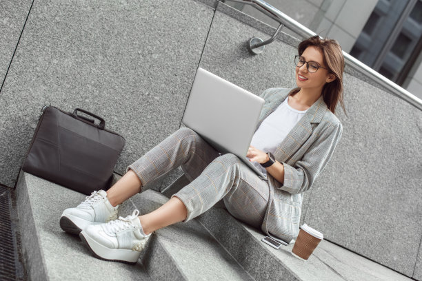 商务休闲装。年轻的女士戴眼镜坐在楼梯上的城市街道与一杯咖啡浏览笔记本电脑快乐图片下载