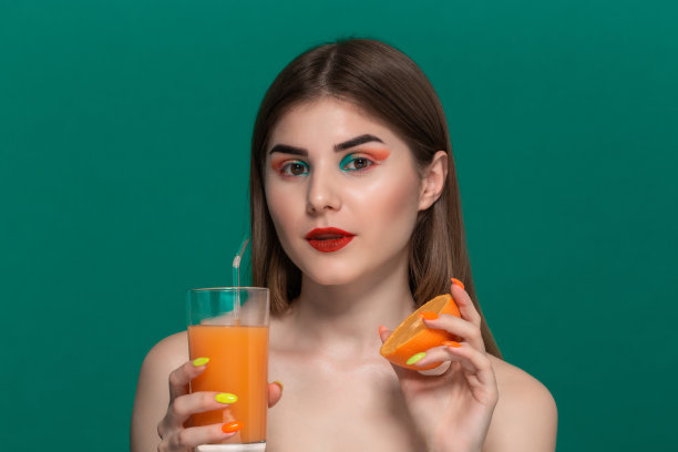 近距离的美丽的年轻女子与明亮的颜色化妆持有橙汁和咬一半的橙子图片下载