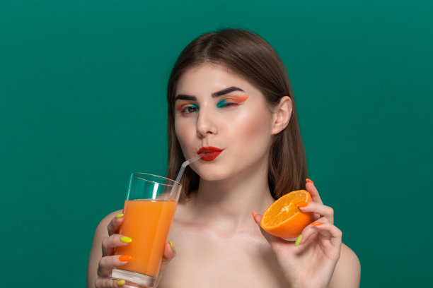 特写美丽的年轻女子与明亮的颜色化妆喝橙汁和持有一半的橙子图片下载