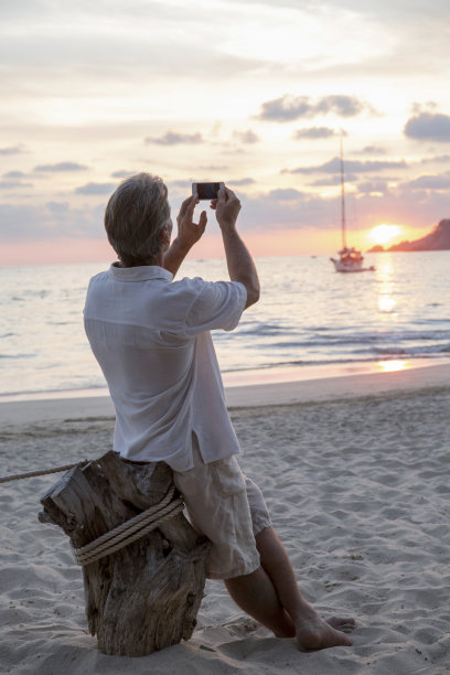 一个成熟的男人在日出的时候用手机在海滩上拍照图片下载