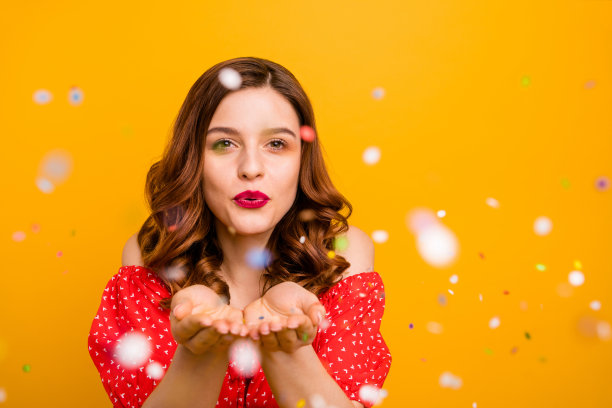 照片惊人的女士吹闪亮的五彩纸屑使童话穿红色连衣裙孤立的黄色背景图片下载