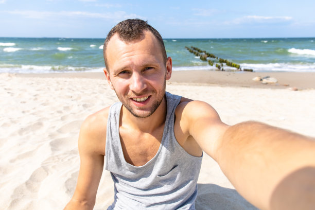 英俊的年轻人坐在海滩上，以大海为背景自拍图片下载