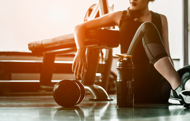 健身女士运动后放松，在健身房旁边放一个乳清蛋白和哑铃。坐在健身房看别处的漂亮年轻女人。图片下载