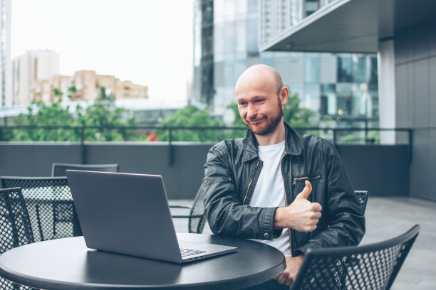 微笑迷人的成年成功秃顶胡子男人，穿着黑色夹克和笔记本电脑在街头咖啡馆的城市图片下载