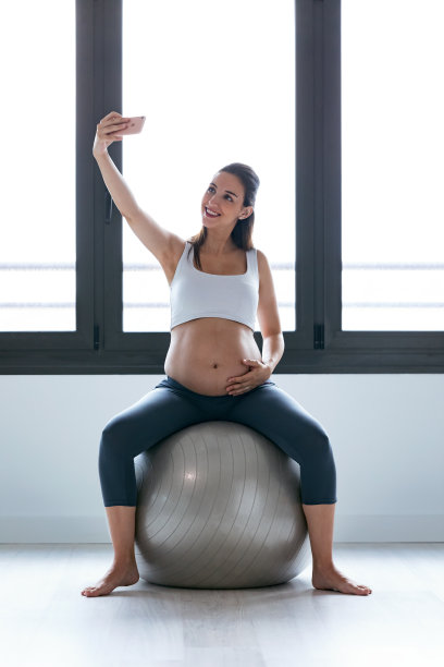 年轻孕妇在家健身球上做放松运动时用手机自拍。图片下载