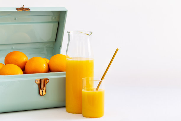 在白色的背景下，一个装满橘子的蓝色金属小提箱，在一个玻璃杯和一个罐子里装着橙汁。图片下载