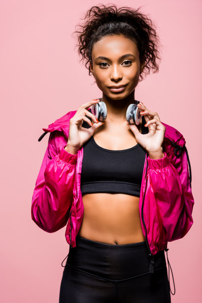 美丽的非洲裔美国女运动员在风衣与耳机看孤立在粉红色的相机图片下载
