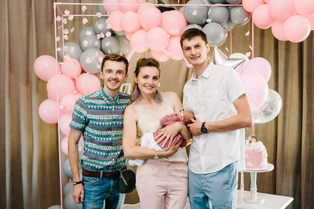 幸福的家庭:母亲，父亲，教父，女儿。父母和朋友的肖像保持在怀里的孩子孩子宝贝女孩，庆祝生日节日派对上的粉红色，灰色的背景与气球。图片下载
