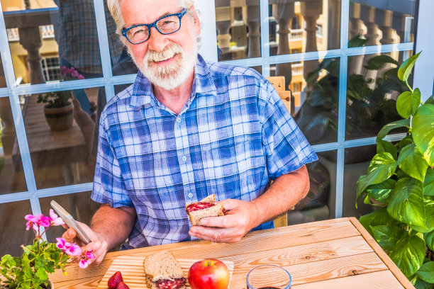 祖父微笑着，满头白发，戴着有色眼镜，吃着意大利腊肠三明治。吃点面包和水果，休息一下。木制的桌子和椅子。手里拿着手机图片下载