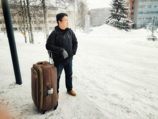 在芬兰的冬天，一名亚裔中国男性游客拿着行李等在被雪覆盖的街道外图片下载