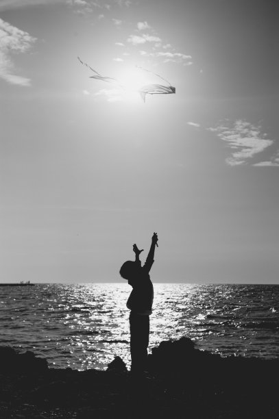 积极自由的小顽皮的孩子的剪影的黑白照片拿着飞翔的风筝，在夏季日落假期上看海岸图片下载