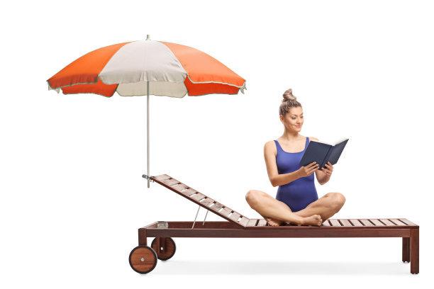 年轻的女人穿着泳衣坐在日光浴床上，拿着雨伞，读着一本书图片下载
