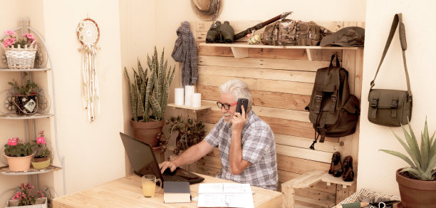 老男人白发大胡子打电话的复古照片。有笔记本电脑和书的木桌。在阳台一角做生意的自由职业者图片下载