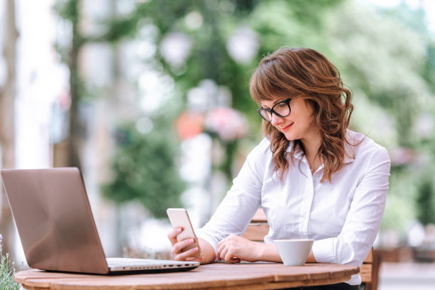 那个穿白衬衫黑裙子的女孩在咖啡馆里一边讲电话一边看笔记本电脑。图片下载