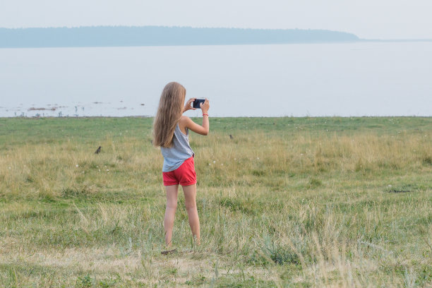 可爱的金发小女孩站在湖边，用智能手机拍风景。图片下载