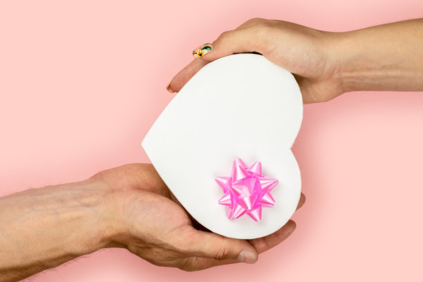 收到一份礼物。男女手握形状心形礼盒粉红色蝴蝶结在粉红色的背景。图片下载