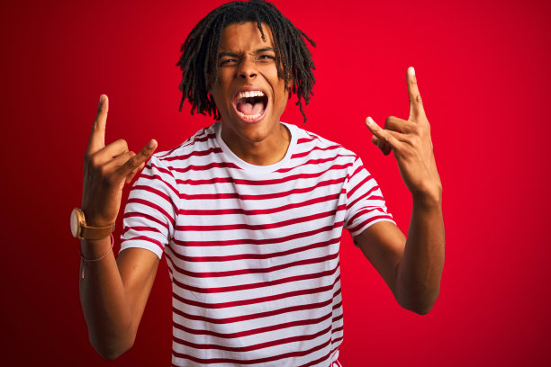 年轻的非洲人与雷鬼发穿着条纹t恤站在孤立的红色背景大喊与疯狂的表情做摇滚符号与手举起。音乐明星。沉重的音乐概念。图片下载