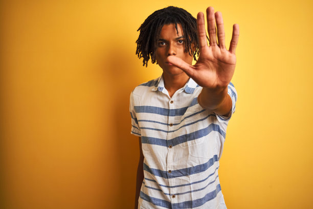 非洲裔美国人与雷鬼发穿着条纹衬衫在孤立的黄色背景做停止唱歌与手掌。警告表情与否定和严肃的手势在脸上。图片下载