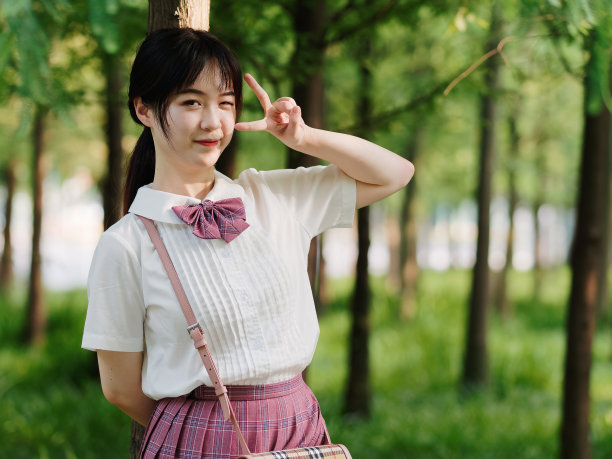 在阳光明媚的夏日森林里，美丽的中国女孩穿着白色衬衫和粉色裙子微笑，用剪刀手势做鬼脸，美丽，生活方式和夏天的概念。素材图片