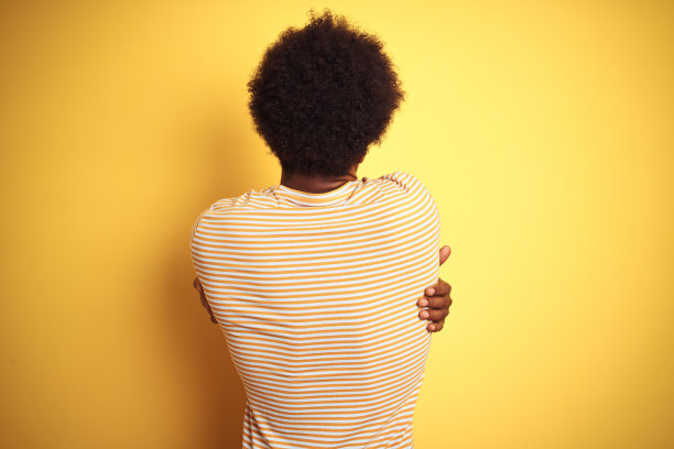 一个留着非洲式发型，穿着条纹t恤的美国人站在孤立的黄色背景上，从背后拥抱着自己快乐和积极。爱自己，关心自己图片下载