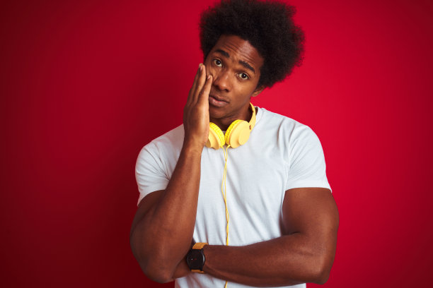 年轻的非洲裔美国人听音乐使用耳机在孤立的红色背景下思考看起来疲倦和厌倦的抑郁问题交叉双臂。图片下载