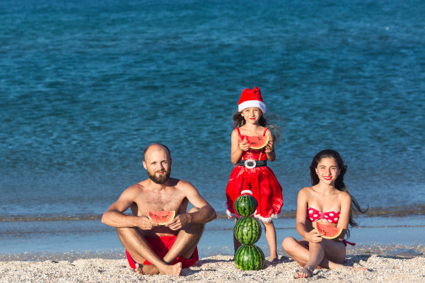 快乐的一家人庆祝夏季圣诞节，父亲和两个女儿在海滩上吃西瓜图片下载