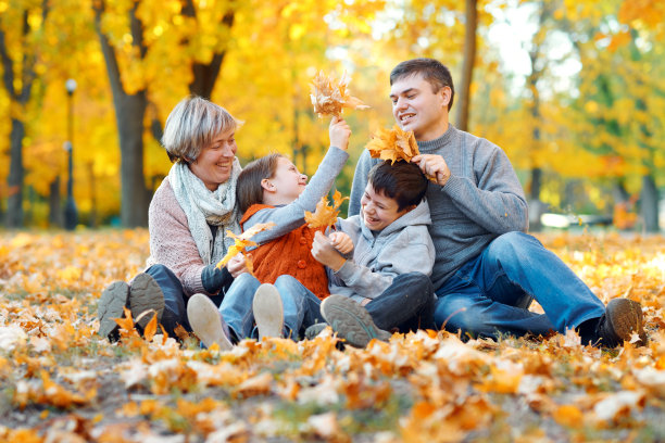 快乐的一家人坐在落叶上，在秋天的城市公园里嬉戏玩耍。孩子和父母一起度过美好的一天。明亮的阳光和树上黄叶，秋天的季节。图片下载
