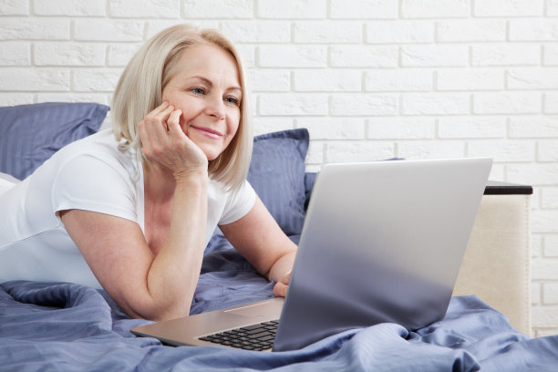 微笑迷人的女人躺在床上使用笔记本电脑在家里在线交流，快乐的女孩打字在电脑上，享受网上购物或在卧室的社交网络聊天。图片下载