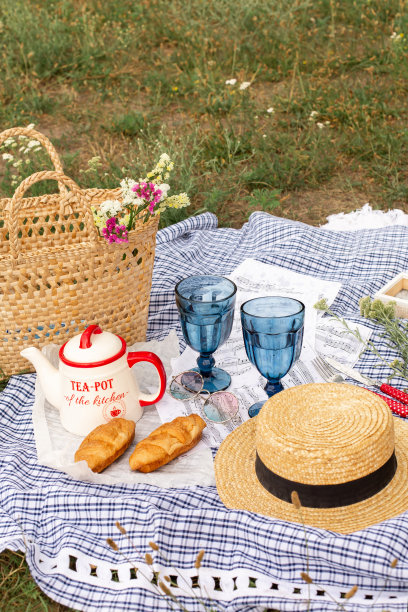 在绿色草坪上的时髦野餐。新鲜的羊角面包，一个茶壶和茶在床罩旁边的柳条女帽。Instagram内容图片下载