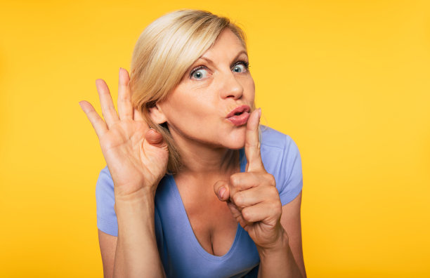 近距离的照片美丽的金发高级女人，而她的手指在她的嘴唇上，显示沉默的手势在黄色的墙壁图片下载